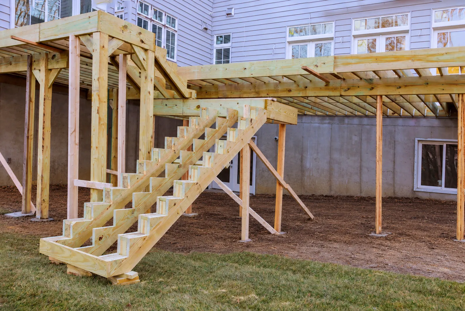 Pressure-treated deck framing and stairs under construction behind a two-story house, shown as a photograph and as a blueprint drawing